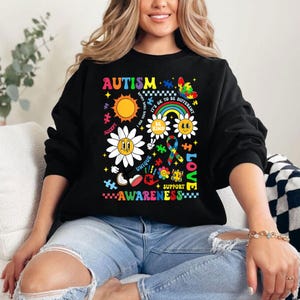 May include: Black sweatshirt with a colorful graphic design featuring the word "AUTISM" and various symbols including puzzle pieces, flowers, and a rainbow. The text "ACCEPT", "UNIQUE", "BE KIND", "IT'S OK TO BE DIFFERENT", "SUPPORT", "LOVE", and "AWARENESS" are also included in the design.
