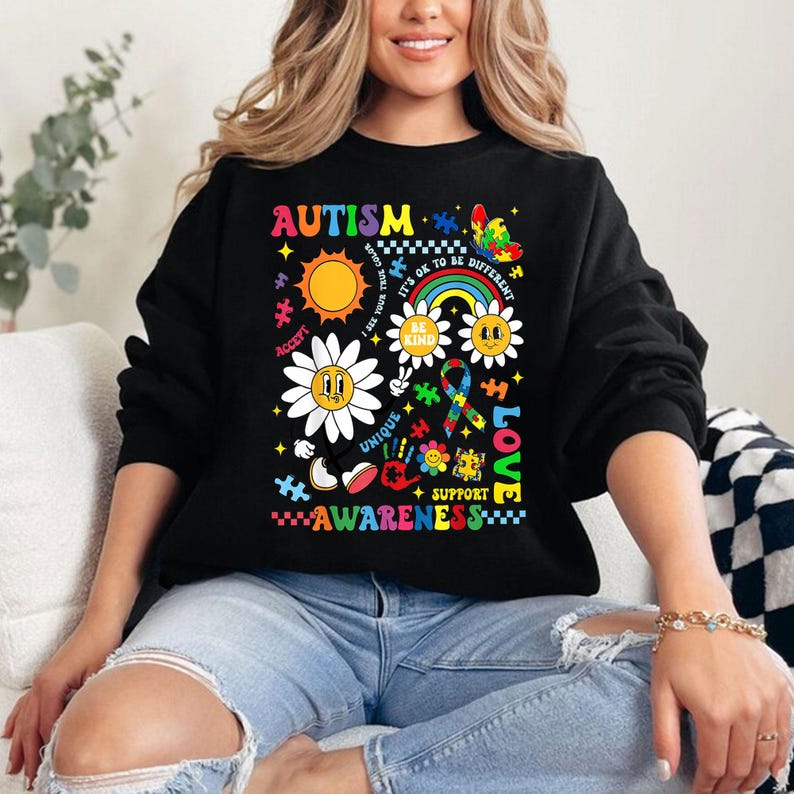 May include: Black sweatshirt with a colorful graphic design featuring the word "AUTISM" and various symbols including puzzle pieces, flowers, and a rainbow. The text "ACCEPT", "UNIQUE", "BE KIND", "IT'S OK TO BE DIFFERENT", "SUPPORT", "LOVE", and "AWARENESS" are also included in the design.