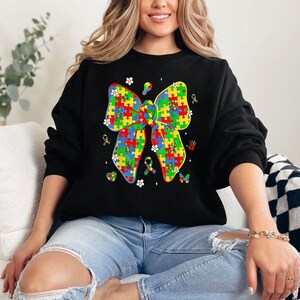 May include: Black sweatshirt with a colorful puzzle piece bow design. The bow is made up of blue, green, yellow, and red puzzle pieces. There are small white flowers, a lightbulb, a handprint, and a butterfly around the bow.