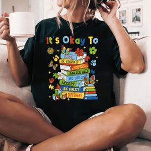 May include: Black t-shirt with a graphic of a stack of colorful books with the text "It's Okay To" and a list of positive affirmations, including "Love", "Be Yourself", "Communicate in Your Own Way", "Ask for Help", "Be Unique", "Accept", "Be First", and "Different".