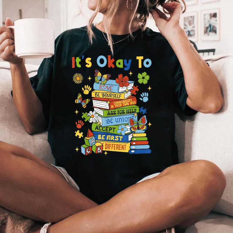 May include: Black t-shirt with a graphic of a stack of colorful books with the text "It's Okay To" and a list of positive affirmations, including "Love", "Be Yourself", "Communicate in Your Own Way", "Ask for Help", "Be Unique", "Accept", "Be First", and "Different".