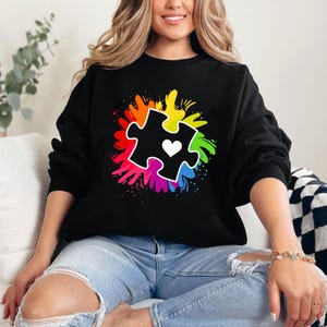May include: Black sweatshirt with a rainbow-colored splatter design featuring a black puzzle piece with a white heart in the center.