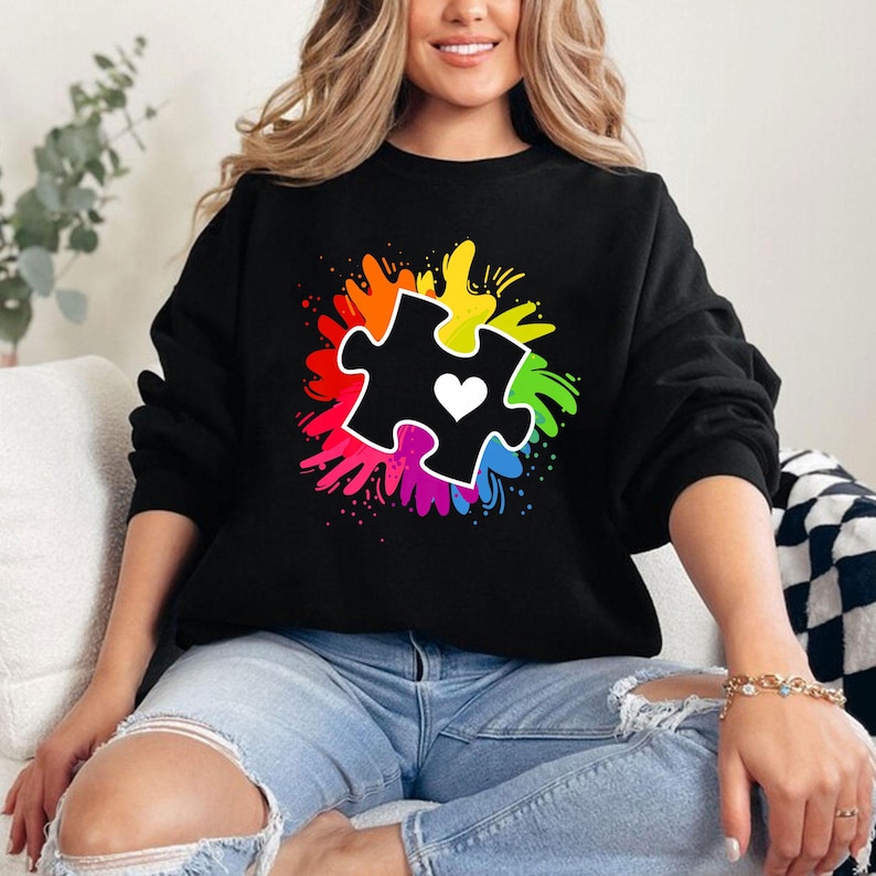 May include: Black sweatshirt with a rainbow-colored splatter design featuring a black puzzle piece with a white heart in the center.