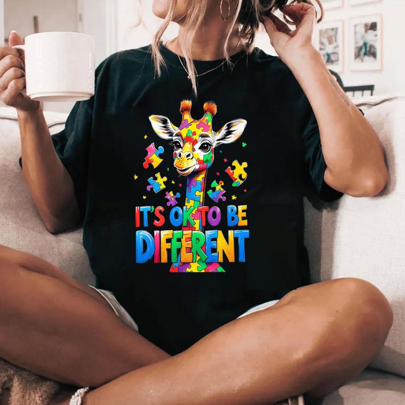 May include: A black t-shirt with a colorful giraffe made of puzzle pieces. The giraffe is surrounded by smaller puzzle pieces. The text "It's ok to be different" is printed below the giraffe.