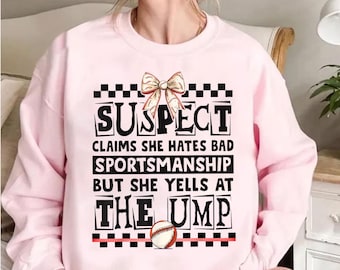 May include: Pink sweatshirt with a black and white checkered design and the text "SUSPECT CLAIMS SHE HATES BAD SPORTSMANSHIP BUT SHE YELLS AT THE UMP" with a baseball graphic.