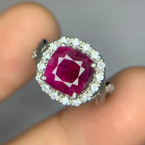Può includere: Un anello d'argento con una grande pietra preziosa di rubino di forma quadrata circondata da un alone di diamanti più piccoli.