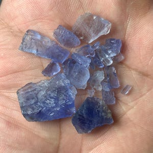 Peut inclure: Un ensemble de cristaux de tanzanite bruts, de tailles et de formes variées. Les pierres précieuses non taillées présentent des nuances de bleu et de violet. Certains cristaux semblent translucides. Les gemmes sont présentées dans une main.
