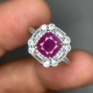 Könnte beinhalten: Ein silberner Ring mit einem großen, quadratisch geschliffenen Rubin im Zentrum, umgeben von einem Halo aus kleineren, runden und baguetteförmigen weißen Edelsteinen. Das Ringband hat ein strukturiertes Design.