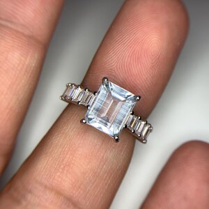 以下が含まれることがあります： エメラルドカットの大きなクリアなアクアマリンと、その周りを囲む小さなバゲットカットのダイヤモンドがセットされたシルバーリングです。