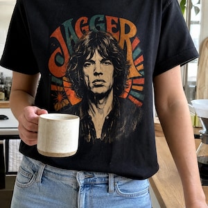 May include: A black short-sleeve t-shirt featuring a graphic print of Mick Jagger with the text "JAGGER" arched above in orange, red, and teal colors. The design includes a sunburst pattern behind the portrait, paired with light blue jeans.