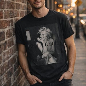 May include: A black short-sleeved t-shirt featuring a square black and white photographic print of Marilyn Monroe with the text MONROE printed in capital letters across the center of the image. The shirt has a crew neckline and a standard fit.
