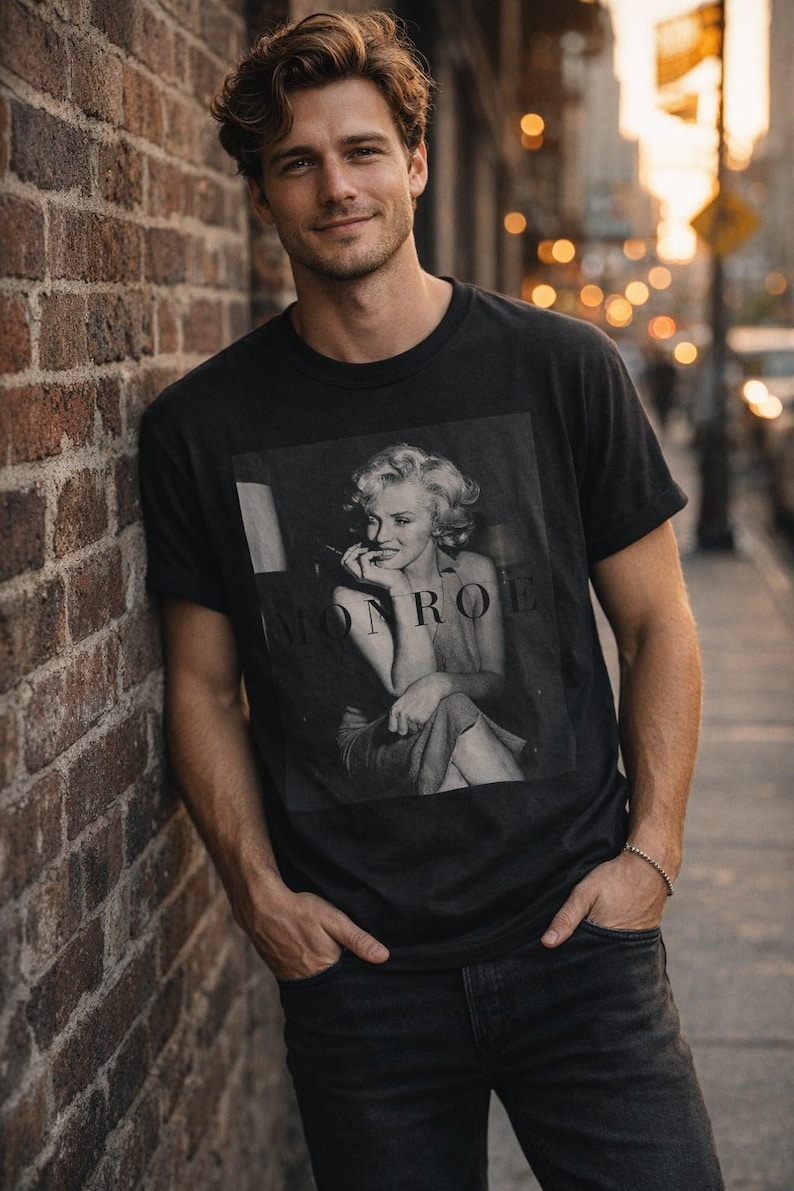 May include: A black short-sleeved t-shirt featuring a square black and white photographic print of Marilyn Monroe with the text MONROE printed in capital letters across the center of the image. The shirt has a crew neckline and a standard fit.