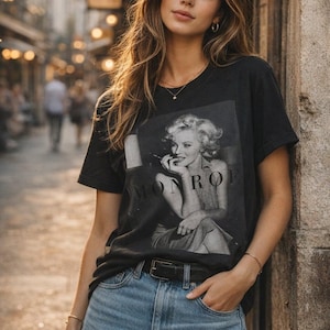 May include: A black short-sleeved t-shirt featuring a monochromatic photographic print of Marilyn Monroe with the text MONROE printed across the center. The shirt is paired with light blue denim jeans and a black leather belt with a silver buckle.