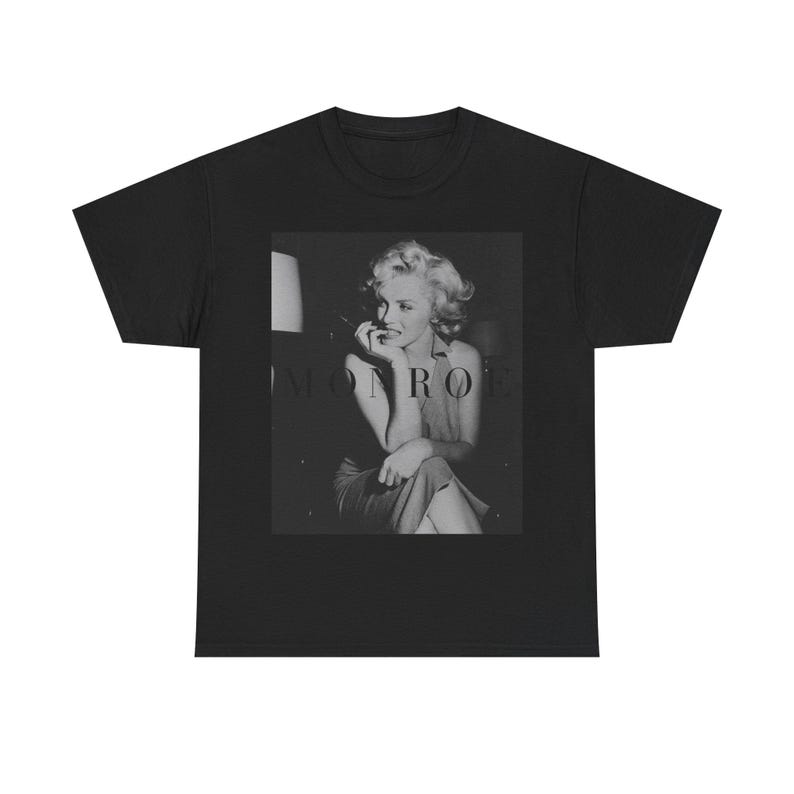 May include: A black short-sleeve t-shirt featuring a centered, rectangular black and white photographic print of Marilyn Monroe. The image shows a portrait of the subject with the text MONROE printed in a dark, subtle font across the center.