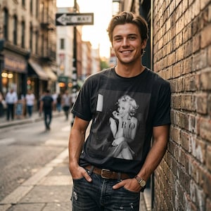 May include: A charcoal gray short-sleeved t-shirt featuring a black and white photographic print of Marilyn Monroe with the text MONROE printed across the center. The shirt is worn with dark blue denim jeans, a brown leather belt, and a wristwatch.