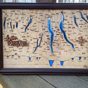 May include: A wooden map of the Finger Lakes region of New York State, with the lakes carved out and painted blue. The map is framed in dark wood and includes the text "New York Finger Lakes".