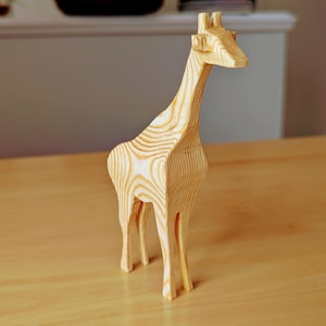 Pode incluir: Uma escultura de girafa de madeira com pescoço longo e pernas finas. A girafa é feita de madeira clara, mostrando os padrões naturais do grão. A escultura está sobre uma superfície de madeira, e o fundo é neutro.