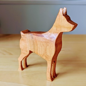 Può includere: Una statuetta di cane in legno scolpita in stile minimalista. Il cane è in piedi su tutte e quattro le zampe e ha un design semplice e geometrico. La statuetta è realizzata in legno marrone chiaro e ha una finitura liscia e lucida.