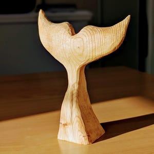 Könnte beinhalten: Eine Holzskulptur einer Walflosse, geschnitzt aus hellem braunem Holz. Die Flosse ist gebogen und hat eine glatte, polierte Oberfläche.