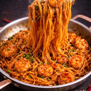 May include: A pan of spaghetti with prawns in a reddish-orange sauce, garnished with spring onions and sesame seeds. Steam rises from the food. Tongs lift a portion of the pasta, showcasing the dish's texture and ingredients.