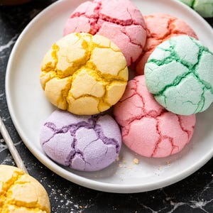 Puede incluir: Un plato blanco contiene varias galletas redondas de superficie agrietada en colores pastel, incluyendo amarillo, rosa, verde claro y lavanda. Las galletas tienen una apariencia texturizada y azucarada con grietas profundas en la parte superior.
