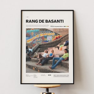 以下が含まれることがあります： 映画のタイトル「Rang De Basanti」が黒字で書かれた額入りのポスター。画像には、カラフルな落書きのある階段に座っている人々が写っています。ポスターは木製のスツールに飾られています。