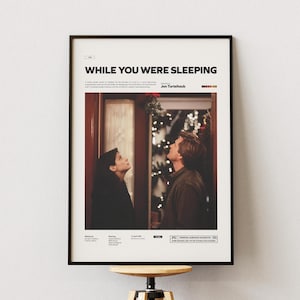 Puede incluir: Un póster enmarcado con el título "While You Were Sleeping" en la parte superior. La imagen muestra a dos personas mirando hacia arriba, con un árbol de Navidad en el fondo. El póster está en un marco negro.