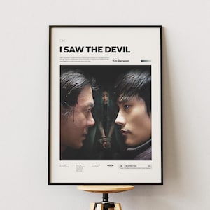 May include: Framed movie poster for "I Saw the Devil" featuring two men facing each other in a dark scene. The poster has a black frame and is mounted on a white wall. The title is at the top, with the director's name below.