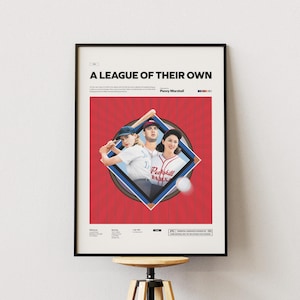 Puede incluir: Póster enmarcado con el título "A League of Their Own" y temática de béisbol. La obra muestra a tres personas con uniformes de béisbol dentro de un rombo, sobre un fondo rojo. El póster está en un marco negro.