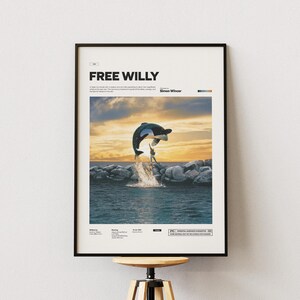 Puede incluir: Póster enmarcado con el título "FREE WILLY". La imagen muestra una orca saltando fuera del agua, con una puesta de sol al fondo. El póster está en un marco negro y se exhibe sobre un taburete de madera.