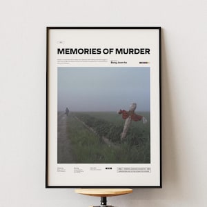 Pode incluir: Pôster emoldurado com o título "MEMORIES OF MURDER" em texto preto. A imagem mostra uma figura em um caminho, um espantalho com tecido vermelho e uma paisagem nebulosa. O pôster está em uma moldura preta.
