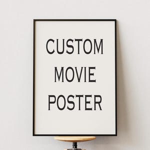 May include: A framed poster with the words "CUSTOM MOVIE POSTER" in a bold, black font. The poster is set against a white background and framed in a black border. The poster is displayed on a wooden stool.