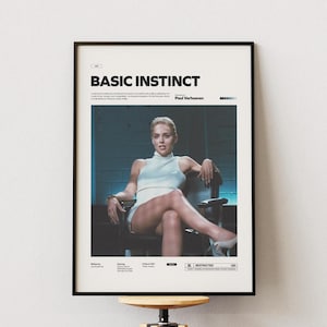 May include: Framed poster featuring the movie title "BASIC INSTINCT" in bold black letters. The image shows a woman seated in a chair, wearing a white outfit. The poster has a black frame and is displayed on a white wall.