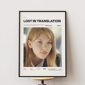 以下が含まれることがあります： 映画「ロスト・イン・トランスレーション」のフレーム入りポスター。金髪の女性の肖像画が描かれています。ポスターは黒いフレームで、白い壁に飾られています。映画のタイトルは上部にあります。