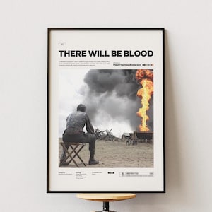 Op de afbeelding: Ingelijste poster voor de film "There Will Be Blood". De poster toont een man die op een kruk zit, wegkijkend, met een brandende oliebron en donkere rook op de achtergrond. De filmtitel staat bovenaan.