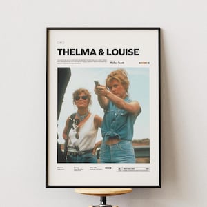 Puede incluir: Póster enmarcado de la película "Thelma & Louise" con el título en la parte superior. La imagen presenta a dos mujeres, una de ellas apuntando con un arma. El póster tiene un marco negro y se muestra en una pared blanca.