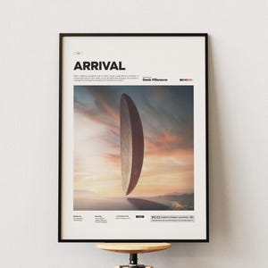 Może przedstawiać: Oprawiony plakat z czarnym napisem "ARRIVAL". Obraz przedstawia duży, pionowy, owalny obiekt zawieszony w powietrzu na tle zachodu słońca. Plakat jest w czarnej ramie.