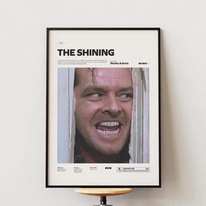 Puede incluir: Póster enmarcado de la película "El Resplandor" con un primer plano del rostro de Jack Nicholson rompiendo una puerta. El título está en la parte superior, con el nombre del director debajo. El póster está en un marco negro.