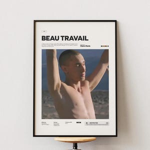Pode incluir: Pôster emoldurado com o título "BEAU TRAVAIL" em letras pretas. O pôster apresenta uma pessoa sem camisa com os braços levantados, em um fundo de céu azul e paisagem desértica. O pôster está em uma moldura preta.