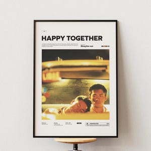 May include: Framed poster for the film "Happy Together." The poster is in a black frame and displays the film's title in bold letters. The image shows two men, one asleep, the other looking ahead. The poster is mounted on a white wall.