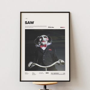 Peut inclure: Affiche encadrée avec le mot "SAW" en haut. L'image montre un personnage du film d'horreur, portant un masque et un nœud papillon, faisant du vélo. L'affiche est dans un cadre noir et est affichée sur un mur blanc.