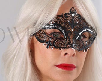 May include: A black filigree mask with rhinestone accents. The mask is worn by a person with blonde hair and red lipstick. The person is wearing a black, one-shoulder sequined top. The text "HEADWINGS ANGELS" is partially visible.