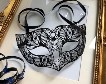 May include: A black, intricately cut masquerade mask with a delicate floral and geometric pattern. The mask is displayed in a gold ornate frame with black ribbons attached. The frame is on a white background.