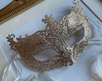 May include: A gold glitter masquerade mask with detailed cut-out patterns and white ribbons. The mask is displayed on a white surface, framed by a gold ornate picture frame, evoking a sense of celebration or theatre.