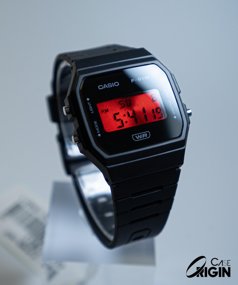 May include: Black Casio F-91W digital watch with a rectangular face and a red digital display showing the time. The watch has a black band and is displayed on a clear stand. The watch face displays the Casio logo.
