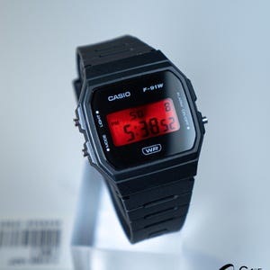 May include: Black Casio F-91W digital watch with a rectangular face and a black resin strap. The red digital display shows the time as 5:38:52 PM. The watch has buttons for light and mode functions. The watch is displayed on a clear stand.
