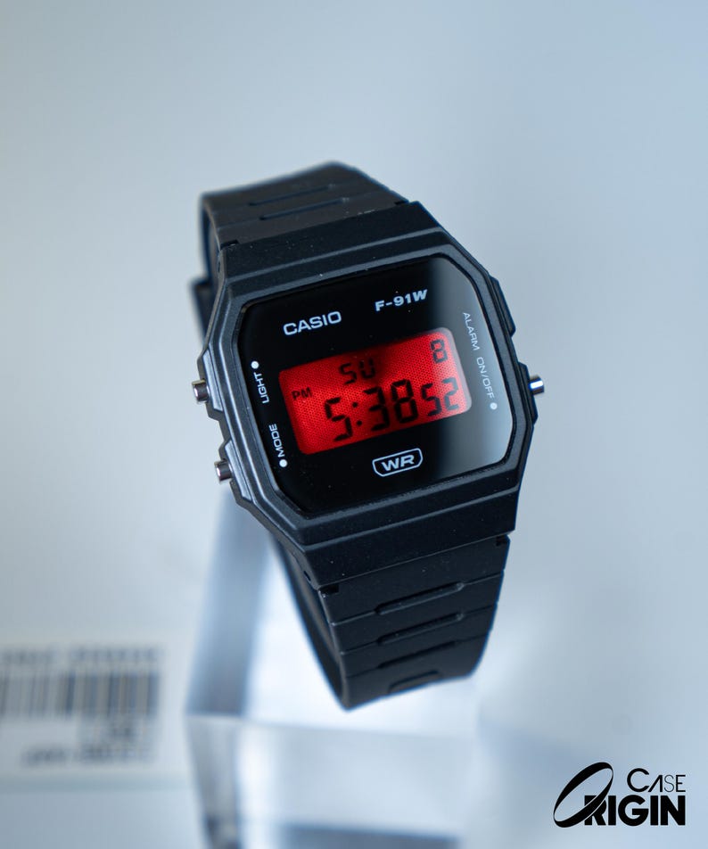 May include: Black Casio F-91W digital watch with a rectangular face and a black resin strap. The red digital display shows the time as 5:38:52 PM. The watch has buttons for light and mode functions. The watch is displayed on a clear stand.