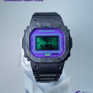 Peut inclure: Une montre noire Casio DW5600 EVA modifiée en fibre de carbone avec un affichage numérique violet et vert. Le cadran affiche l'heure et le texte "EVANGELION" et "TOKYO-3". La montre a un bracelet en fibre de carbone noir.
