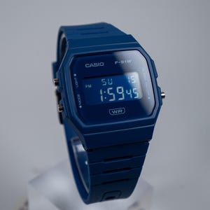 Può includere: Un orologio da polso digitale blu scuro con cassa rettangolare e cinturino in resina abbinato. Il quadrante mostra la scritta CASIO F-91W, WR e un orario digitale di 1:59:45 con l'indicatore del giorno SU 15. Sono visibili tre pulsanti argentati.