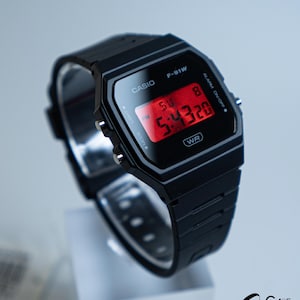 May include: A black Casio F-91W digital watch with a rectangular face and a black resin strap. The red digital display shows the time. The watch has the text "CASIO" and "F-91W" printed on the face.